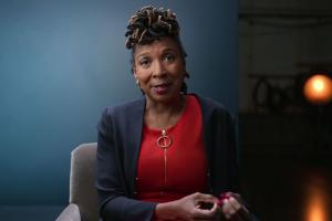 KIMBERLÉ CRENSHAW, Professor, UCLA and Columbia Schools of Law. She is an African-American woman, wearing a red blouse under a navy blue blazer; she is seated against a blue backdrop.