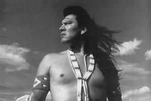 A Native American man, wearing traditional Indigenous accoutrements, stands in the middle of a plain, looking to his right. Courtesy of TBS