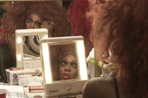 BeBe Zahara Benet is a Black drag queen preparing for a show in her dressing room in Minneapolis. She is wearing a very large brown wig and false eyelashes, while looking into a mirror. From Emily Branham’s ‘Being BeBe.’ Courtesy of the filmmaker.