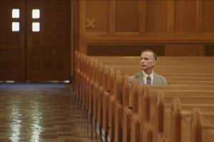 Michael Sandridge is a white man in a grey suit sitting alone inside a church. From Robert Greene’s “Procession.” Courtesy of Netflix.