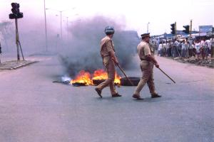 Tires burning in the middle of the road, two Indian police officers are walking by with batons in hand and crowd watching on the side of the road