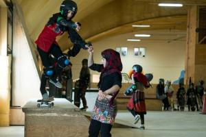 A young Afghani girl in a checkered headscarf helps her friend on a skateboarding ramp. Her friend is wearing skateboarding gear. They’re inside a training room with other young skateboarders. Image from Carol Dysinger’s ‘Learning to Skateboard in a Warzone (If You're a Girl).’ Courtesy of the filmmaking team.
