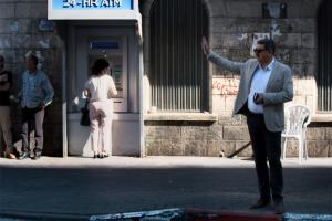 Musa Hadid, the mayor of Ramallah is a middle-aged man with a moustache. He is waving to someone and is wearing sunglasses, and is wearing a grey jacket with a shirt and trousers. There are passersby and an ATM machine behind him. Image from David Osit’s 'Mayor.' Courtesy of David Osit and POV.