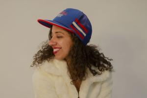 A photo of filmmaker Justine Armen, a mixed-race woman with dark brown curly hair wearing a blue and red baseball cap and white coat, looking to the side and smiling.