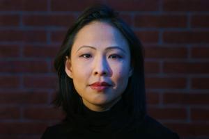An Asian woman of Malaysian Chinese origin with shoulder-length black hair. She is wearing a black turtleneck & in front of a brick wall.