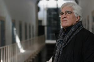 A Spanish man with white hair and glasses is staring into the distance from inside of a prison