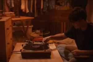 A young guy plays a record player in a small, cramped bedroom.