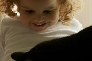 a black cat plays on the lap of a White female toddler.