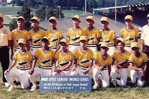 The 1992 Little League World Series Champion team from the Philippines pose in front of the baseball stadium in yellow jerseys and hats. Their coaches stand on either side of the players. 