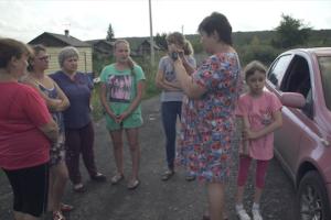 Homemaker-turned-journalist Natalia Zubkova in a blue and pink flowered dress, interviews five women with her cell phone in a rural village as her young daughter, in pink shirt and capri pants, waits nearby.
