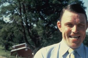A Middle-aged White man smiles in a blue button-up and yellow tie in front of a rural set of houses.