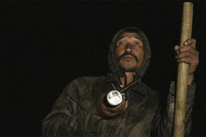 A man holds a wooden staff in the dark.