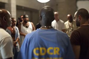 A Black Male Folsom inmate stands in a blue shirt in front of a diverse group of male prisoners.