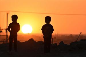 Two silhouetted children stand before the setting sun.