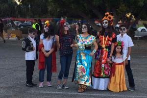 A group of Mexican-American women and children in Oaxacan dress and Dia de los Muertos make-up stand in a parking lot.