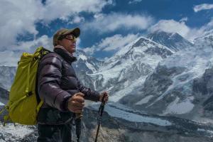 Renowned climber Minga Tsiri Sherpa stands holding walking poles in his hands and wearing a backpack as he looks out towards the Himalayan mountains which are also seen behind him in the distance. 