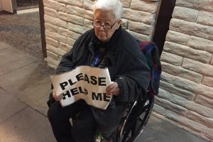 an elderly asian individual in a wheelchair holds a sign reading "please help me"