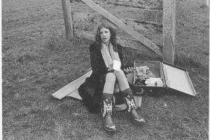 Young woman in early 70's fashion sits on a picnic blanket next to an open suitcase with a notepad in her hand.