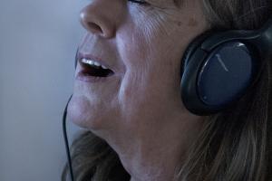A middle-aged white woman with blue headphones closes her eyes as she adjusts a voicebox on her neck.