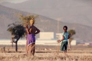 An Ethiopian farmer wearing a yellow headscarf and flowery dress and her daughter in ponytail wearing a blue T-shirt and green skirt stand in dry teff field looking into the distance against a hazy background of a massive industrial zone. 