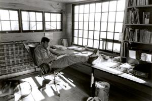 a black and white image of Wendell Berry reading a book in a chair with feet kicked up on table