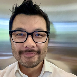 A man with glasses, spikey black hair and a black mustache smiles at the camera in a selfie-style photo. He wears a white collared shirt.