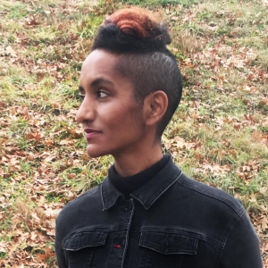 A woman with her head shaved on the side and mohawk style hairdo looks to the side. She wears a dark blue collard shirt buttoned to the top. Behind her is an autumnal grass.