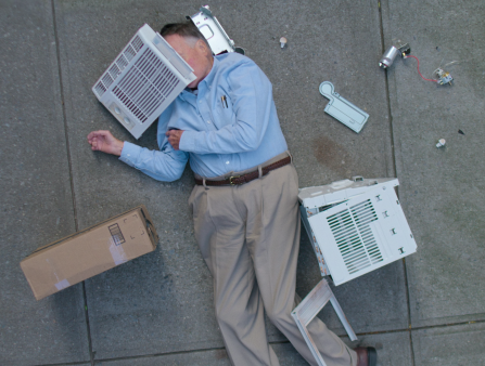 Still from Dick Johnson is Dead. A person on their back on the ground wearing a blue button up shirt, brown belt, and khaki pants, next to a brown box on one side and on the other side a broken air conditioner with part of it covering their face.