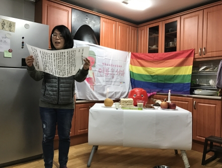 In a kitchen with a rainbow flag and cakes on a table, a woman reads from a scroll.