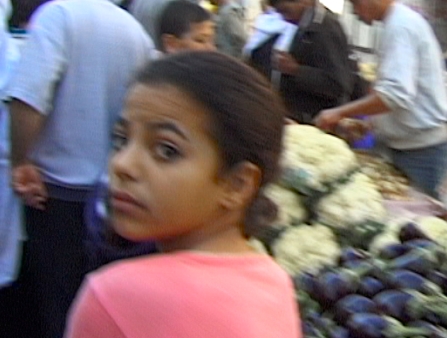 A young girl in a pink shirt looks over her shoulders. Behind her is a crowd of men.