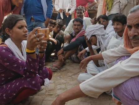 A young woman interviews an older man on her phone, while many other gather round them.