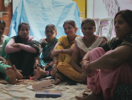 A group of women sit during a meeting. Out of the right side of a frame, is a person holding a camera.