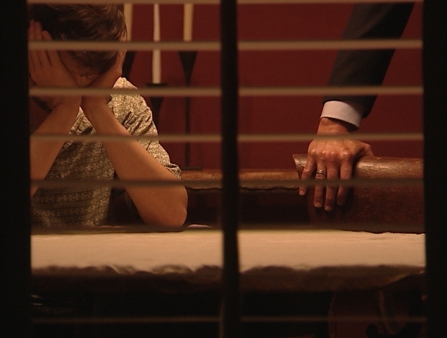Video still of a young man with his face in his hands. Besides him, an arm grips the back of a couch.