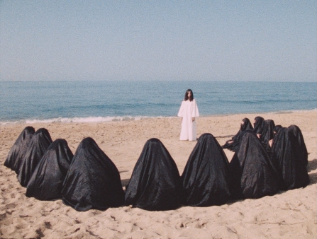On a beach, a lone woman in white stands in the background, looking at a semicircle of seated women in black burqas.