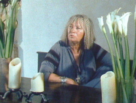 A woman with a bob sits at a dining table with floral arrangements.