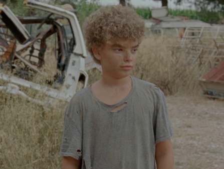 A young boy with curly blond hair stands in the middle of a car junkyard.
