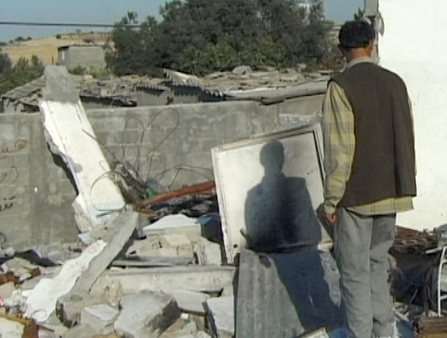 A man wearing a vest looks at a pile of destroyed belongings.