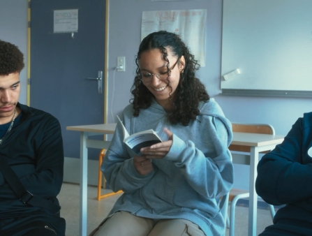 Three young people look at a book, while sitting in a classroom.