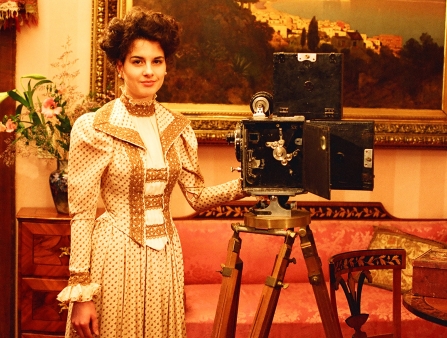 A woman in a long-sleeved dress stands behind an old film camera.