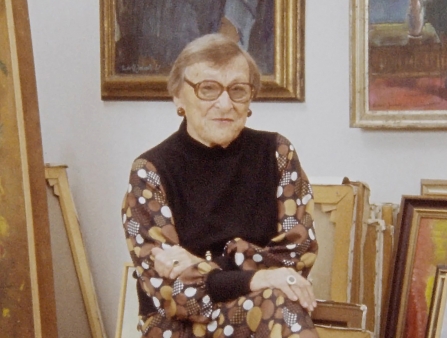A woman sits in an art studio filled with paintings.
