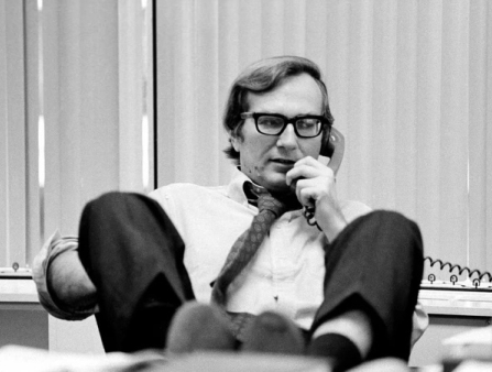 A white man on a phone call, while propping his legs up on his desk.