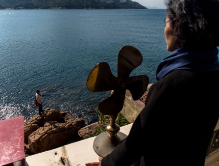A woman looks over a ledge at a man standing on the rocks by a large expanse of water.