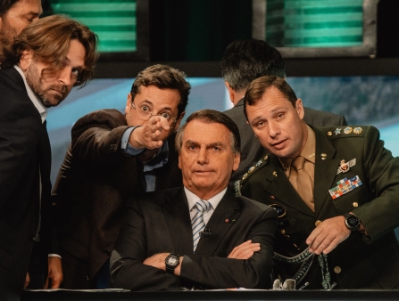 An angered older man in a suit and tie with crossed arms is surrounded by four men, one of whom is pointing at the camera.