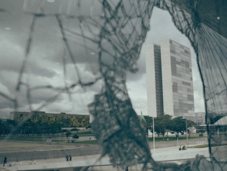 The hole in a broken window frames a tall white building against a stormy sky.