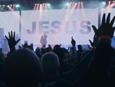 At a religious revival meeting, a man preaches to the crowd. There is a giant screen that reads: Jesus.