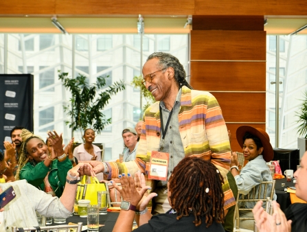 A Black man with graying hair and glasses stands while being applauded by his peers.