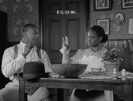 A young couple, dressed in clothes from the early 20th century, sits at a table. The woman is signing: "F-L-O-W."