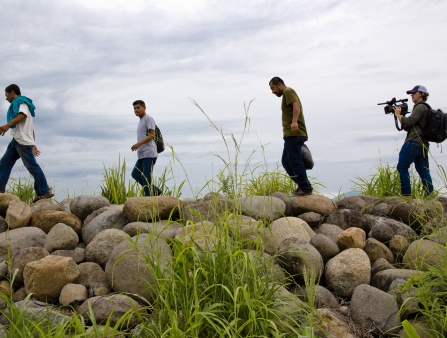 A man holding a camera films a group of three other young men walking along a rocky bank.
