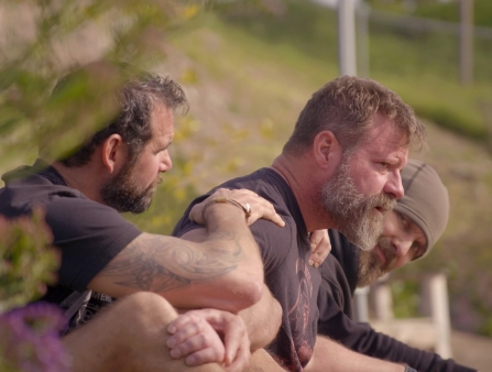 Film still of three grizzled white men sitting on a hillside.