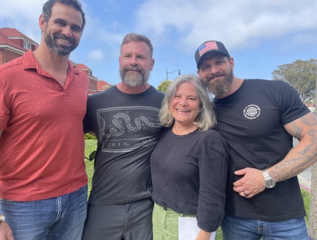 Under blue skies, four white people embrace for the camera.